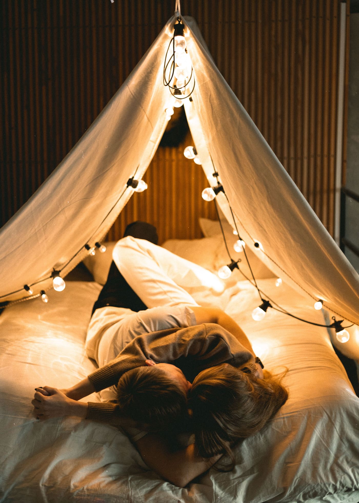 A couple embracing in bed under warm string lights, faces close together and turned away from camera